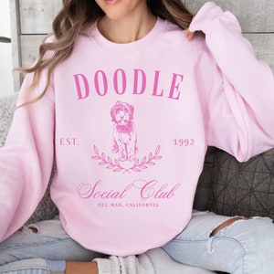 Puede incluir: Sudadera rosa con el texto "Doodle Social Club" en blanco, con una ilustraci&oacute;n de un perro doodle y el texto "Est. 1992" y "Del Mar, California".