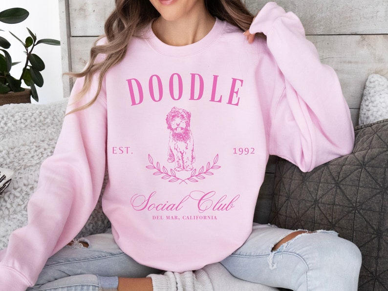 Puede incluir: Sudadera rosa con el texto "Doodle Social Club" en blanco, con una ilustraci&oacute;n de un perro doodle y el texto "Est. 1992" y "Del Mar, California".