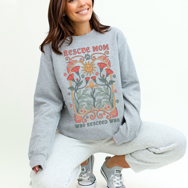 Puede incluir: Sudadera gris con un dise&ntilde;o gr&aacute;fico de un sol, flores y el texto "Rescue Mom Who Rescued Who".