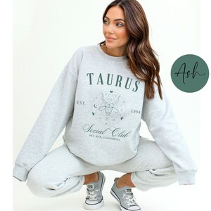 Puede incluir: Una sudadera gris con un dise&ntilde;o gr&aacute;fico verde que dice "Taurus Social Club Est. 1994 Del Mar, California".