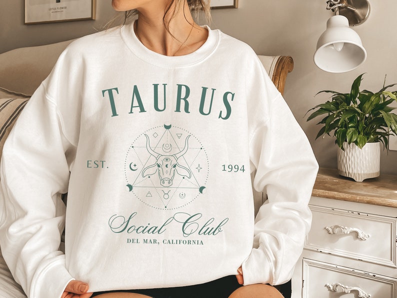 Puede incluir: Sudadera blanca con un dise&ntilde;o gr&aacute;fico verde que presenta la palabra "TAURUS" y una cabeza de toro estilizada con un patr&oacute;n geom&eacute;trico. El texto "EST. 1994" y "Social Club" tambi&eacute;n est&aacute;n incluidos en el dise&ntilde;o.