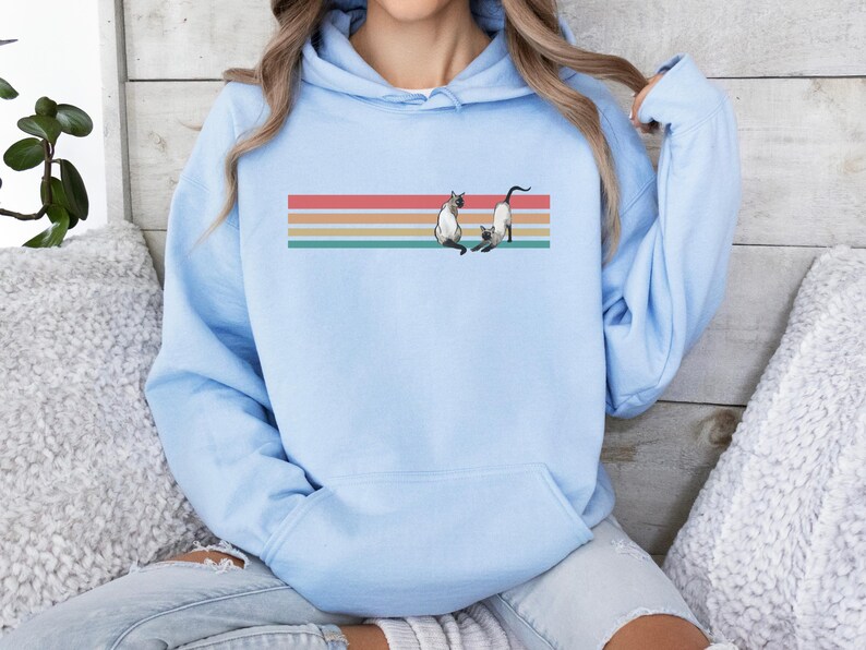 May include: Light blue hoodie featuring a graphic of two Siamese cats on a horizontal striped design in red, orange, yellow, and teal. The hoodie has a front pocket and a drawstring hood.