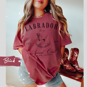 May include: A maroon t-shirt with a black graphic design featuring a Labrador Retriever, the text "Labrador Roxy Social Club Est. 2024 El Mar, California", and a wreath of leaves.