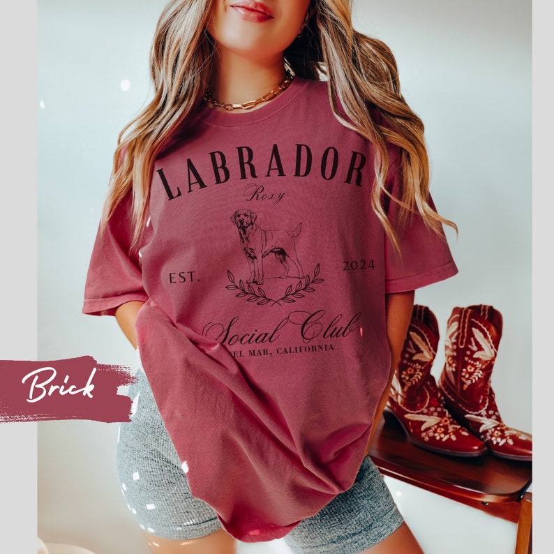 May include: A maroon t-shirt with a black graphic design featuring a Labrador Retriever, the text "Labrador Roxy Social Club Est. 2024 El Mar, California", and a wreath of leaves.