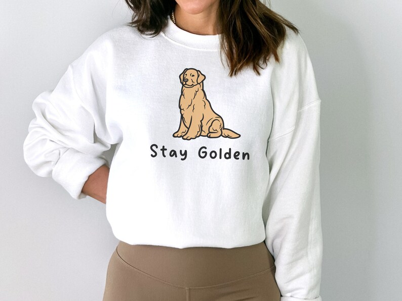 以下が含まれることがあります： 前面に卡通のゴールデンレトリバーが座っているイラストと「Stay Golden」という文字がプリントされた白いスウェットシャツです。