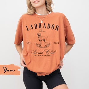 May include: A burnt orange t-shirt with a black graphic design featuring a Labrador Retriever, the text "Labrador", "Roxy", "Est. 2024", and "Social Club Del Mar, California".