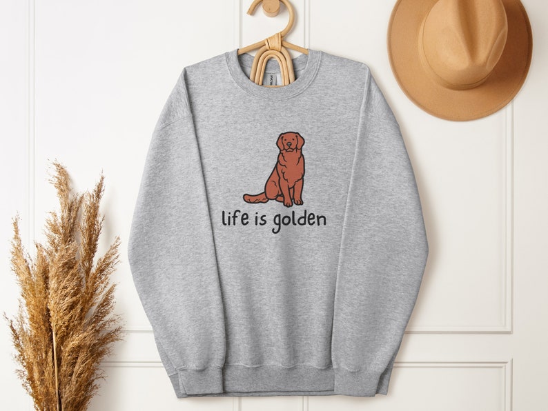 Op de afbeelding: Een grijs sweatshirt met een illustratie van een bruine golden retriever en de tekst "life is golden".