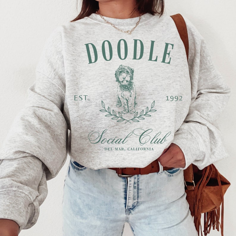 Puede incluir: Una sudadera gris con un gr&aacute;fico de perro doodle y el texto "Doodle Social Club Est. 1992 Del Mar, California".