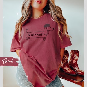 May include: A brick red t-shirt with a graphic of a dachshund wearing a beret and the text "oui-ner".