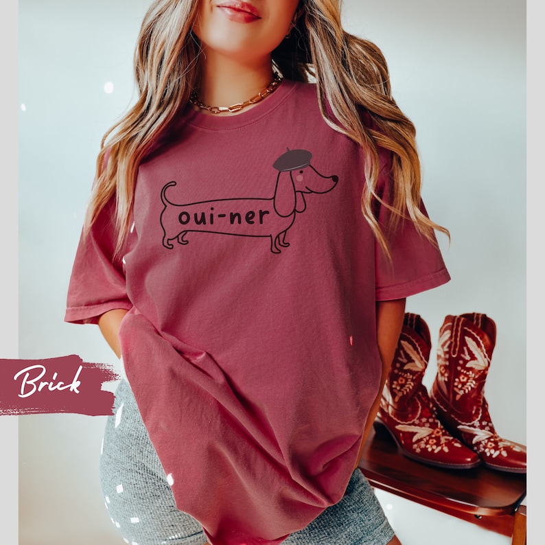 May include: A brick red t-shirt with a graphic of a dachshund wearing a beret and the text "oui-ner".
