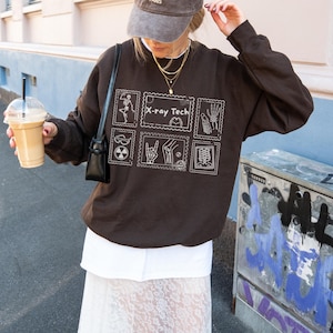 May include: A dark brown sweatshirt with white graphic illustrations and the text "X-ray Tech". The design includes images of bones, hands, and other medical symbols. The sweatshirt is worn with a white skirt and a gray cap.
