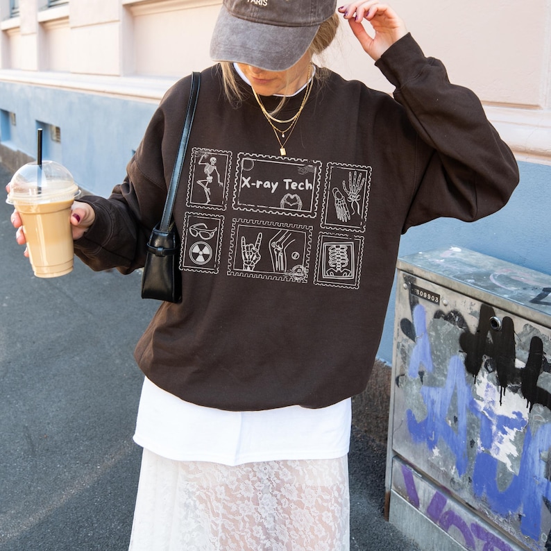 May include: A dark brown sweatshirt with white graphic illustrations and the text "X-ray Tech". The design includes images of bones, hands, and other medical symbols. The sweatshirt is worn with a white skirt and a gray cap.