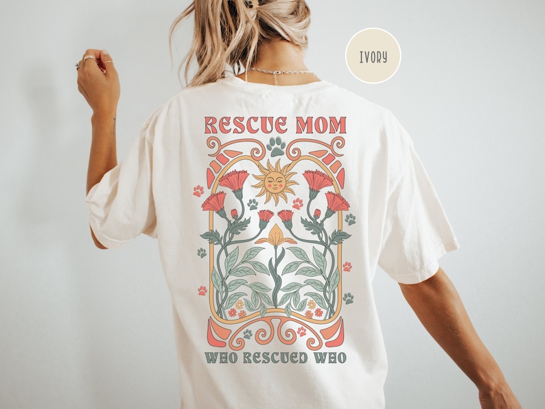 May include: Ivory t-shirt with a graphic design of a sun, flowers, and paw prints. The text "Rescue Mom Who Rescued Who" is printed on the shirt.