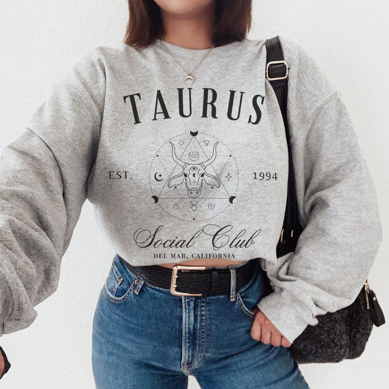 Puede incluir: Sudadera gris con un dise&ntilde;o gr&aacute;fico negro que presenta el signo del zod&iacute;aco Tauro, una cabeza de toro dentro de un c&iacute;rculo con formas geom&eacute;tricas y el texto "Taurus Social Club Est. 1994 Del Mar, California".