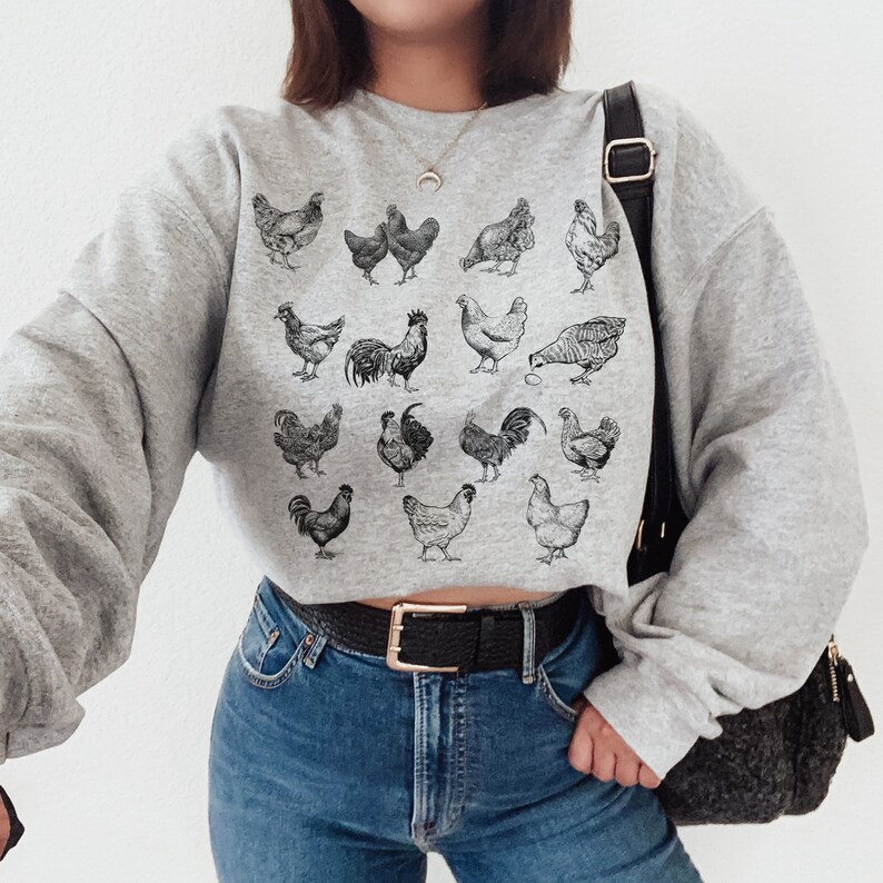 May include: A gray sweatshirt with a black and white illustration of various chickens. The sweatshirt has a cropped fit and is being worn by a person with long brown hair.