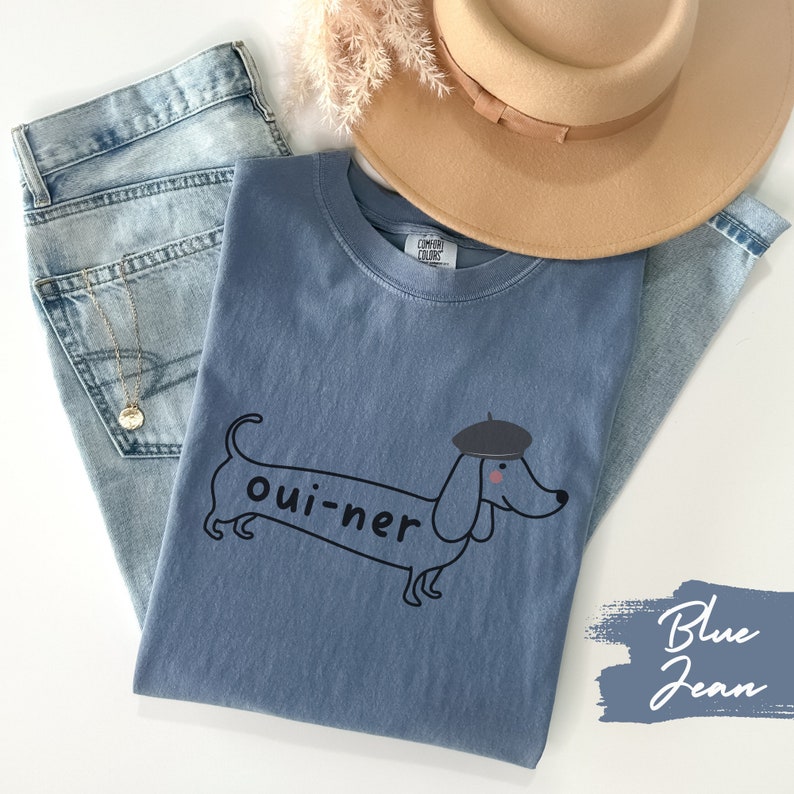 May include: A blue jean t-shirt with a cartoon dachshund wearing a beret and the text "oui-ner".