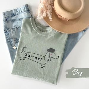 May include: A light green t-shirt with a black and white illustration of a dachshund wearing a beret. The dachshund is holding a banner that says "oui-ner". The t-shirt is laying on a light blue denim background with a brown hat and a sprig of dried flowers.