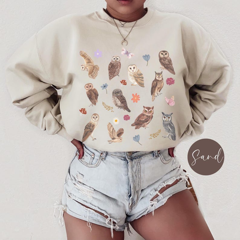 May include: A beige sweatshirt with a pattern of owls, flowers, and mushrooms. The sweatshirt has long sleeves and a crew neck.