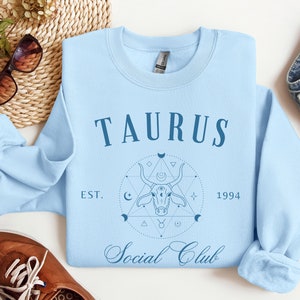 Puede incluir: Sudadera azul claro con un dise&ntilde;o gr&aacute;fico de una cabeza de toro en un c&iacute;rculo con estrellas y lunas. El texto "Taurus Social Club" est&aacute; impreso en la sudadera. El texto "EST. 1994" est&aacute; impreso debajo del gr&aacute;fico.