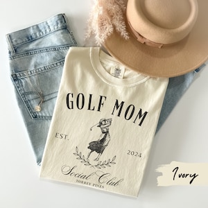 K&ouml;nnte beinhalten: Ein T-Shirt in Elfenbein mit schwarzem Text, der "GOLF MOM EST. 2024 Social Club TORREY PINES" lautet, und einer Schwarzwei&szlig;illustration einer Frau, die Golf spielt.