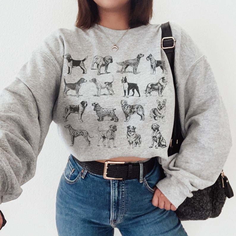 May include: A gray cropped sweatshirt with a black and white print of various dog breeds. The sweatshirt is worn by a person wearing blue jeans and a black belt.