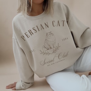 May include: A beige sweatshirt with a brown graphic print of a Persian cat, the text "Persian Cat", "Est. 1984", "Social Club", and "Del Mar, California".