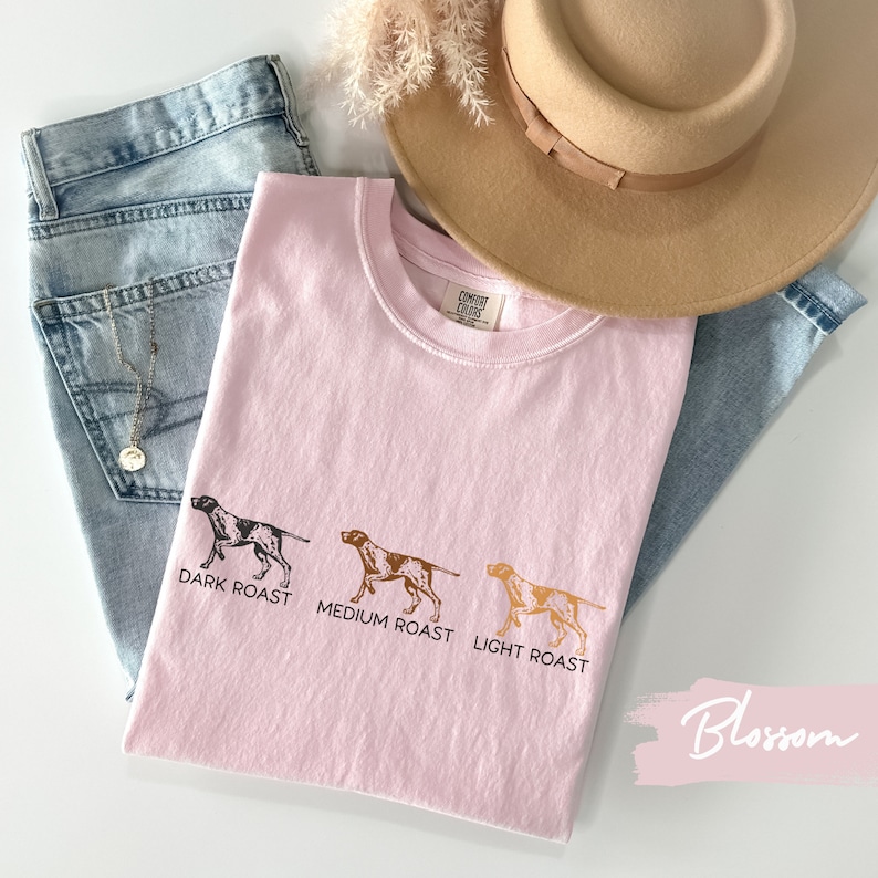 Op de afbeelding: Roze T-shirt met een afbeelding van drie honden in verschillende kleuren en de tekst "DARK ROAST, MEDIUM ROAST, LIGHT ROAST". Het shirt ligt op een lichtblauwe denim achtergrond met een bruine hoed en een gouden ketting.