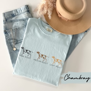 Op de afbeelding: Lichtblauw Chambray T-shirt met een afbeelding van drie jachthonden die verschillende koffieroasters vertegenwoordigen: donker geroosterd, medium geroosterd en licht geroosterd.