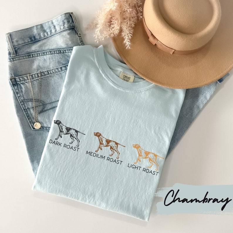 Op de afbeelding: Lichtblauw Chambray T-shirt met een afbeelding van drie jachthonden die verschillende koffieroasters vertegenwoordigen: donker geroosterd, medium geroosterd en licht geroosterd.