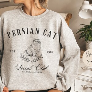 May include: A grey sweatshirt with a black graphic design featuring a Persian cat, the text "Persian Cat Social Club", and the dates "Est. 1984".