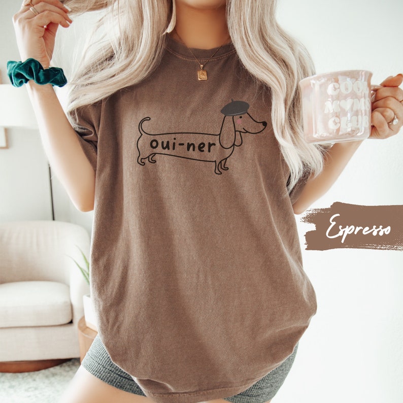 May include: A brown t-shirt with a cartoon dachshund wearing a beret and the text "oui-ner" printed on the front. The shirt is being modeled by a person holding a mug that says "COOP MY WINE CLUB".