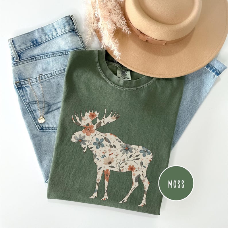May include: A moss green t-shirt with a floral moose design, paired with light wash denim jeans, a tan hat, and a gold necklace. A green circle with the word "MOSS" is in the lower right corner.