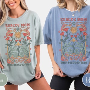 May include: Two women wearing oversized t-shirts with a vintage-style graphic design. The text on the shirts reads "Rescue Mom Who Rescued Who". The shirts are available in two colors: bay and blue jean.