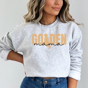 May include: A gray sweatshirt with the text "GOLDEN mama" in black and tan. The word "GOLDEN" is in large, bold letters, and the word "mama" is in a smaller, cursive font. A silhouette of a golden retriever is incorporated into the design.