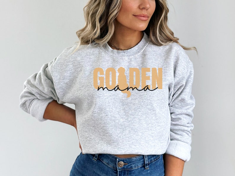 May include: A gray sweatshirt with the text "GOLDEN mama" in black and tan. The word "GOLDEN" is in large, bold letters, and the word "mama" is in a smaller, cursive font. A silhouette of a golden retriever is incorporated into the design.
