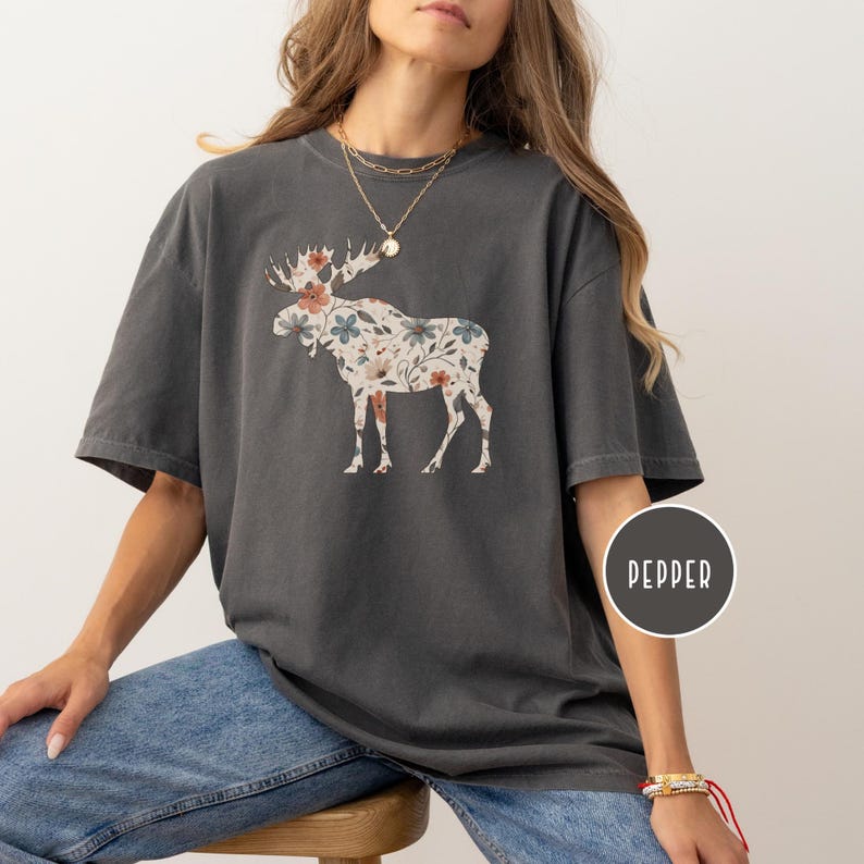 May include: Dark grey t-shirt with a floral moose design. The moose is patterned with pink, blue, and white flowers. The shirt has short sleeves and a relaxed fit, suitable for everyday wear.