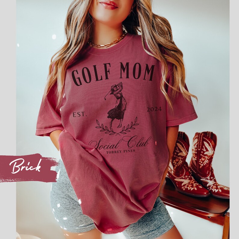 K&ouml;nnte beinhalten: Ein maronfarbenes T-Shirt mit dem Text "GOLF MOM EST. 2024 Social Club TORREY PINES" und einer Grafik einer Frau, die Golf spielt.