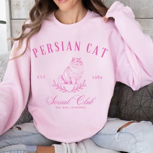 May include: A pink sweatshirt with the text "PERSIAN CAT" in pink lettering. The sweatshirt also features a drawing of a Persian cat, the text "EST. 1984", and the text "Social Club" with the text "DEL MAR, CALIFORNIA" underneath.