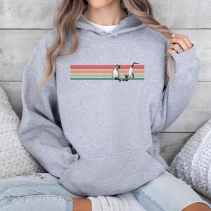 May include: A light gray hoodie featuring a graphic of two Siamese cats on a horizontal striped design in red, orange, yellow, and teal. The hoodie has a front pocket and a hood with a drawstring.