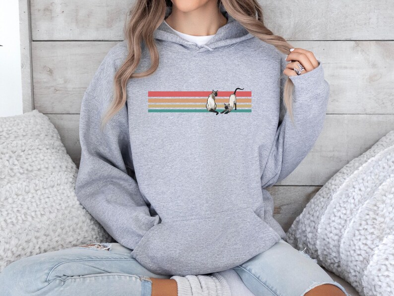 May include: A light gray hoodie featuring a graphic of two Siamese cats on a horizontal striped design in red, orange, yellow, and teal. The hoodie has a front pocket and a hood with a drawstring.