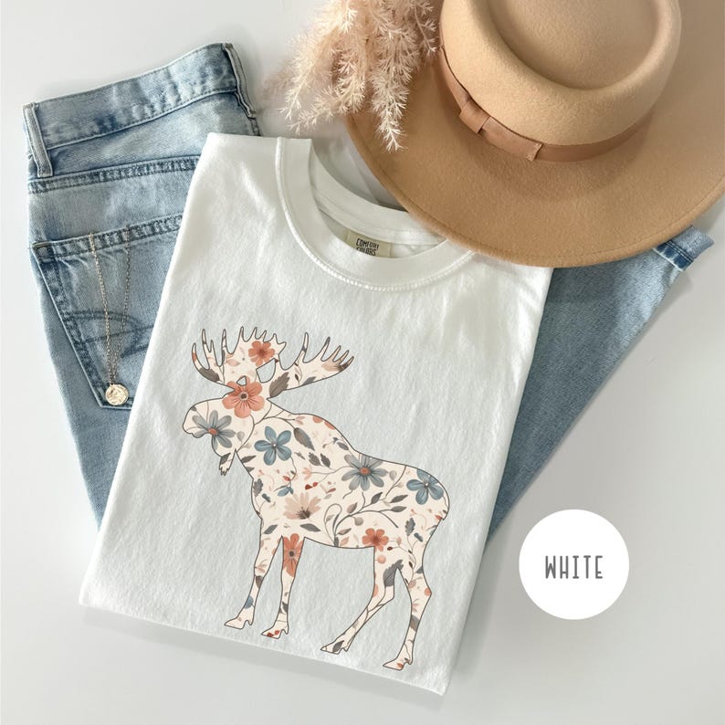 May include: White t-shirt with a floral moose design in peach, blue, and cream. The shirt is styled with light blue jeans and a tan hat. A gold necklace is visible. The word "WHITE" is in a white circle.
