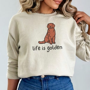 Op de afbeelding: Een beige sweatshirt met een cartoon golden retriever en de tekst "life is golden".
