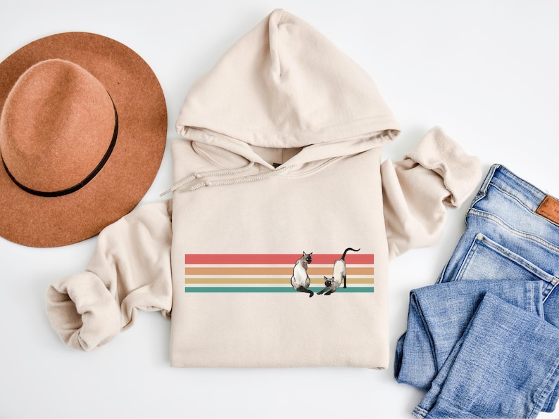 May include: Beige hoodie with a retro design featuring a horizontal stripe in red, orange, yellow, and teal. The design includes two Siamese cats. A brown hat and blue jeans are also in the image.