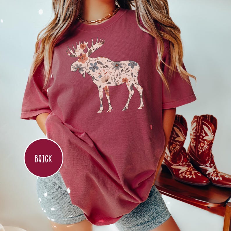 May include: A brick-red t-shirt with a floral moose design. The moose is filled with a pattern of flowers in shades of pink, grey, and white. The word "BRICK" is printed on a circular label in the lower left corner.