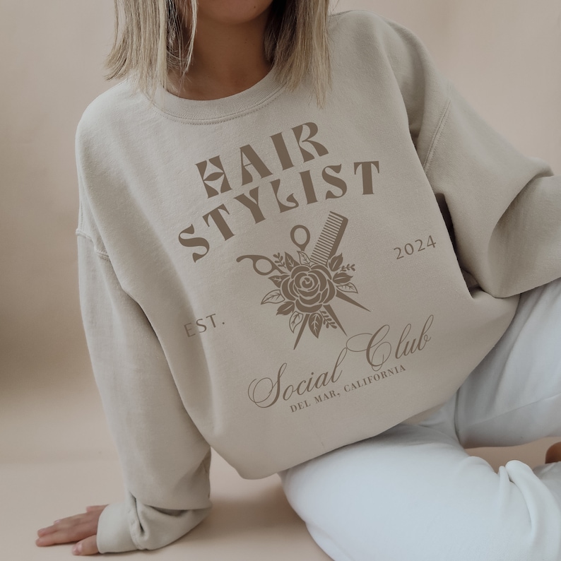 K&ouml;nnte beinhalten: Ein beiger Pullover mit dem Text "HAIR STYLIST" in braunen Buchstaben. Der Pullover zeigt au&szlig;erdem eine Grafik von einer Schere und einem Kamm, die &uuml;ber einer Rose gekreuzt sind, mit dem Text "EST. 2024 Social Club DEL MAR, CALIFORNIA".