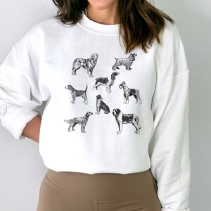 K&ouml;nnte beinhalten: Wei&szlig;er Pullover mit einer Schwarzwei&szlig;illustration verschiedener Hunderassen. Die Hunde sind alle im Profil und schauen nach rechts. Der Pullover hat einen Rundhalsausschnitt und lange &Auml;rmel.