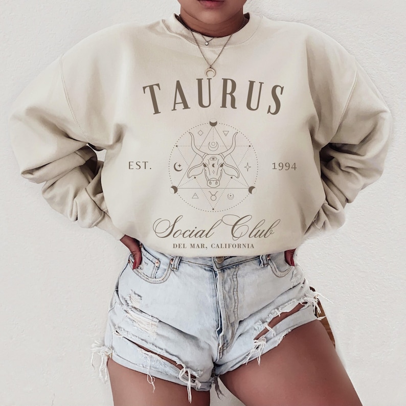 Puede incluir: Una sudadera beige con el texto "TAURUS EST. 1994 Social Club DEL MAR, CALIFORNIA" y un gr&aacute;fico de una cabeza de toro dentro de un c&iacute;rculo con s&iacute;mbolos astrol&oacute;gicos.