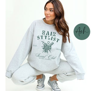 K&ouml;nnte beinhalten: Ein grauer Pullover mit dem Text "Hair Stylist Social Club Est. 2024 Del Mar, California" und einer Grafik von einer Schere, einem Kamm und einer Rose.