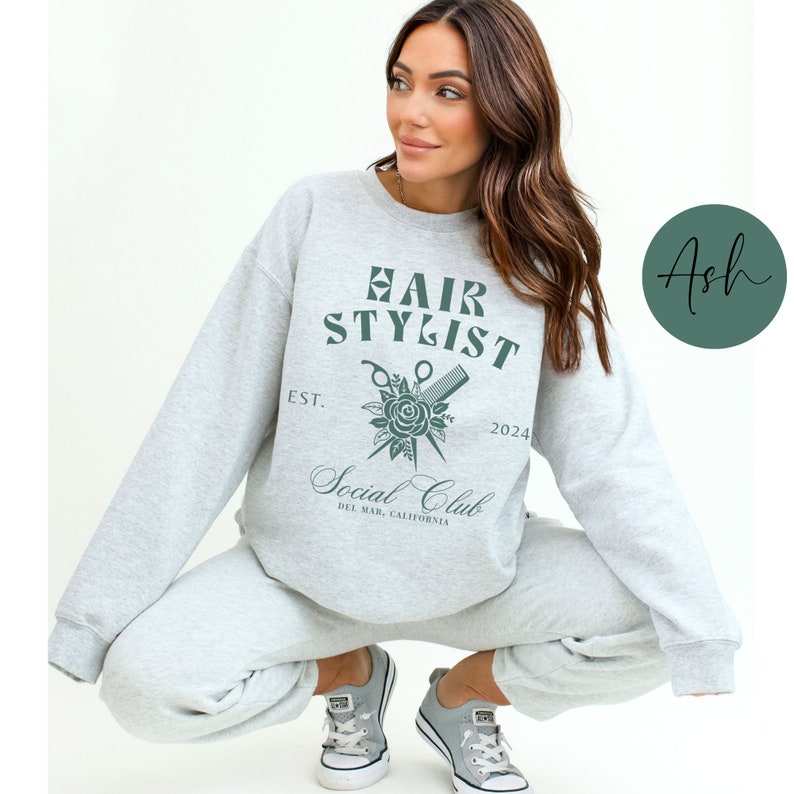 K&ouml;nnte beinhalten: Ein grauer Pullover mit dem Text "Hair Stylist Social Club Est. 2024 Del Mar, California" und einer Grafik von einer Schere, einem Kamm und einer Rose.