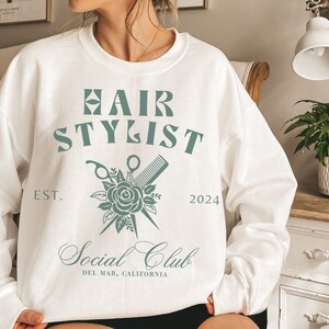 K&ouml;nnte beinhalten: Wei&szlig;er Pullover mit einem gr&uuml;nen Grafikdesign, auf dem "Hair Stylist" mit einer Rose, einer Schere und einem Kamm steht. Der Text darunter lautet "Social Club" und "Del Mar, Kalifornien".